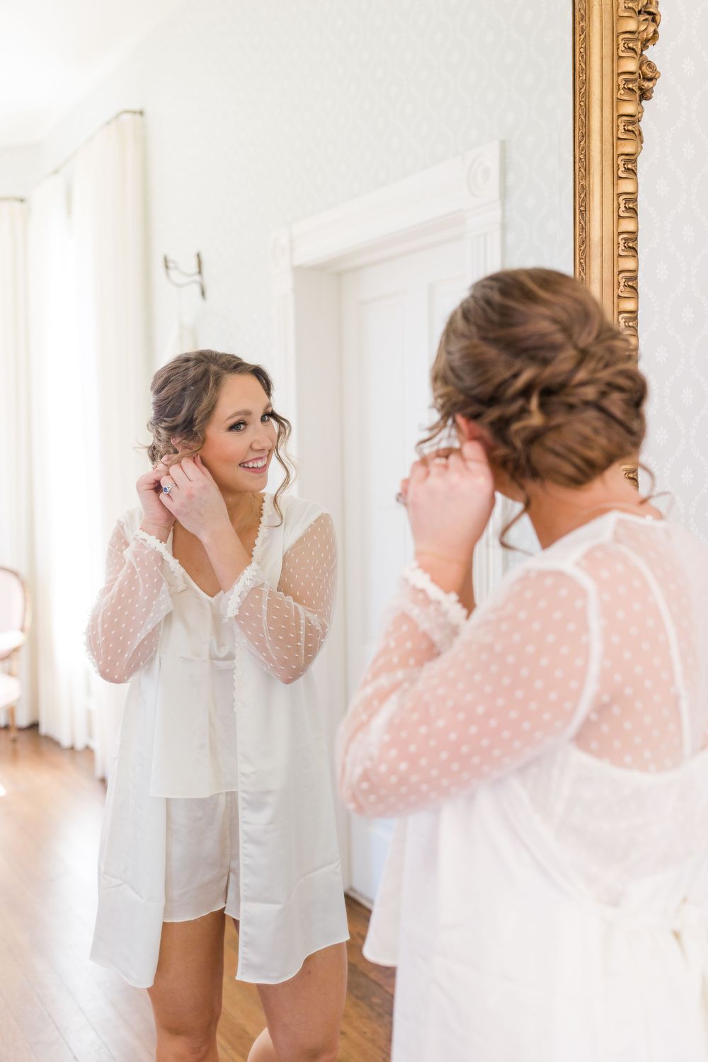 Bride in white getting ready robe admiring reflection while putting on earrings.