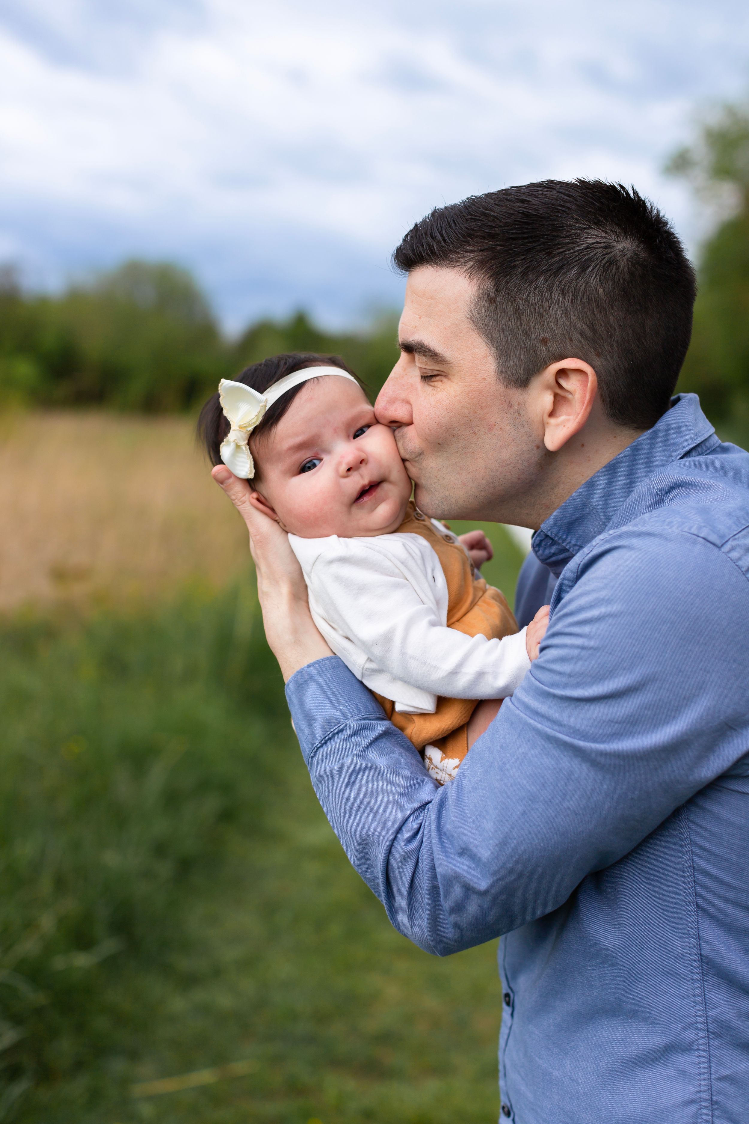 A tender kiss shared with a baby wearing white against a natural outdoor setting.