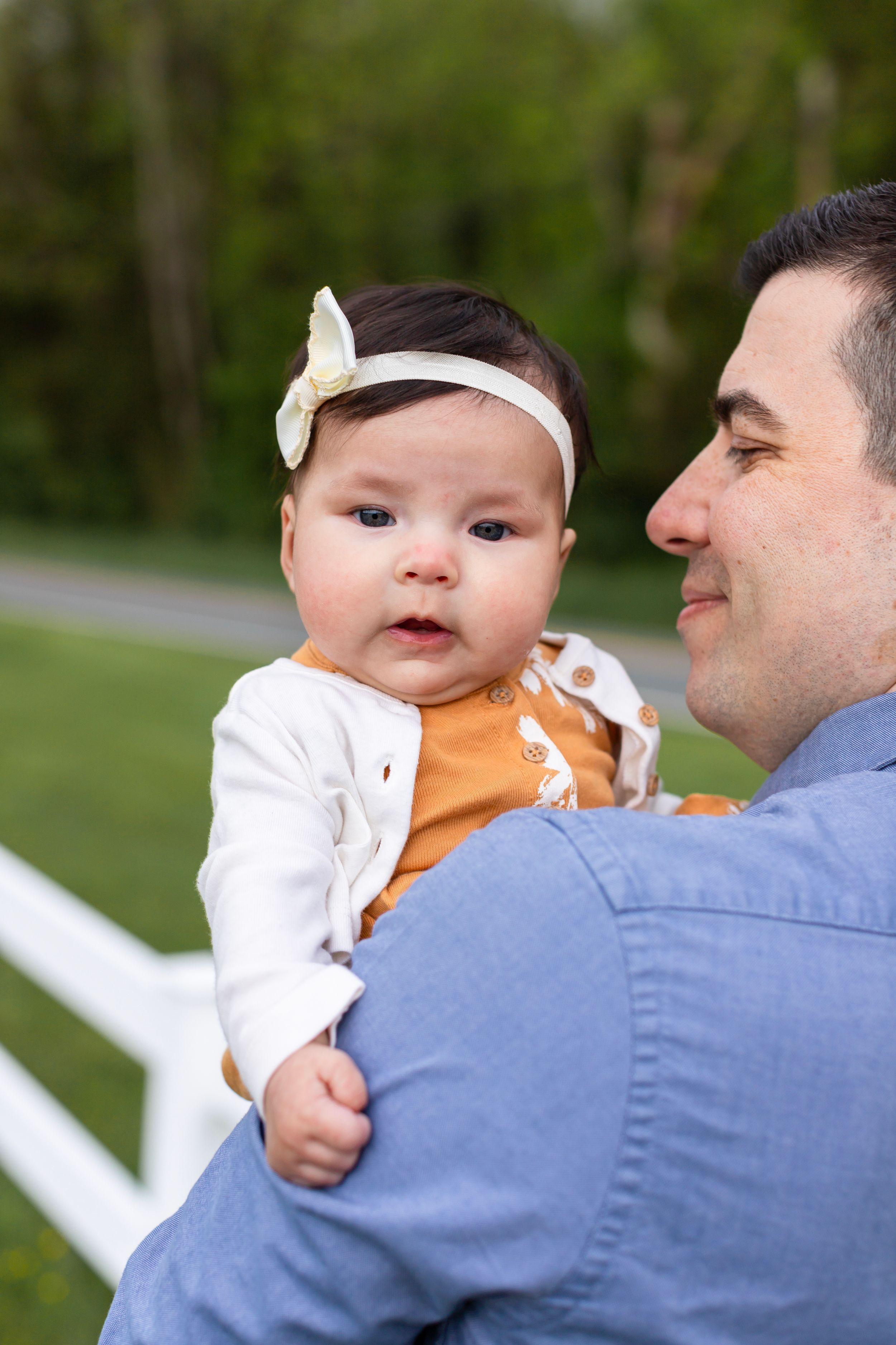 A baby wearing a white bow headband is held against a backdrop of white fencing.