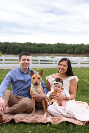 Outdoor family portrait on picnic blanket with dog in field with white fence.