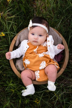 Baby in orange romper with white headband sits in wooden bowl surrounded by wildflowers.
