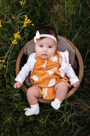 Baby wearing orange romper and white bow sits in wooden bowl among yellow flowers.