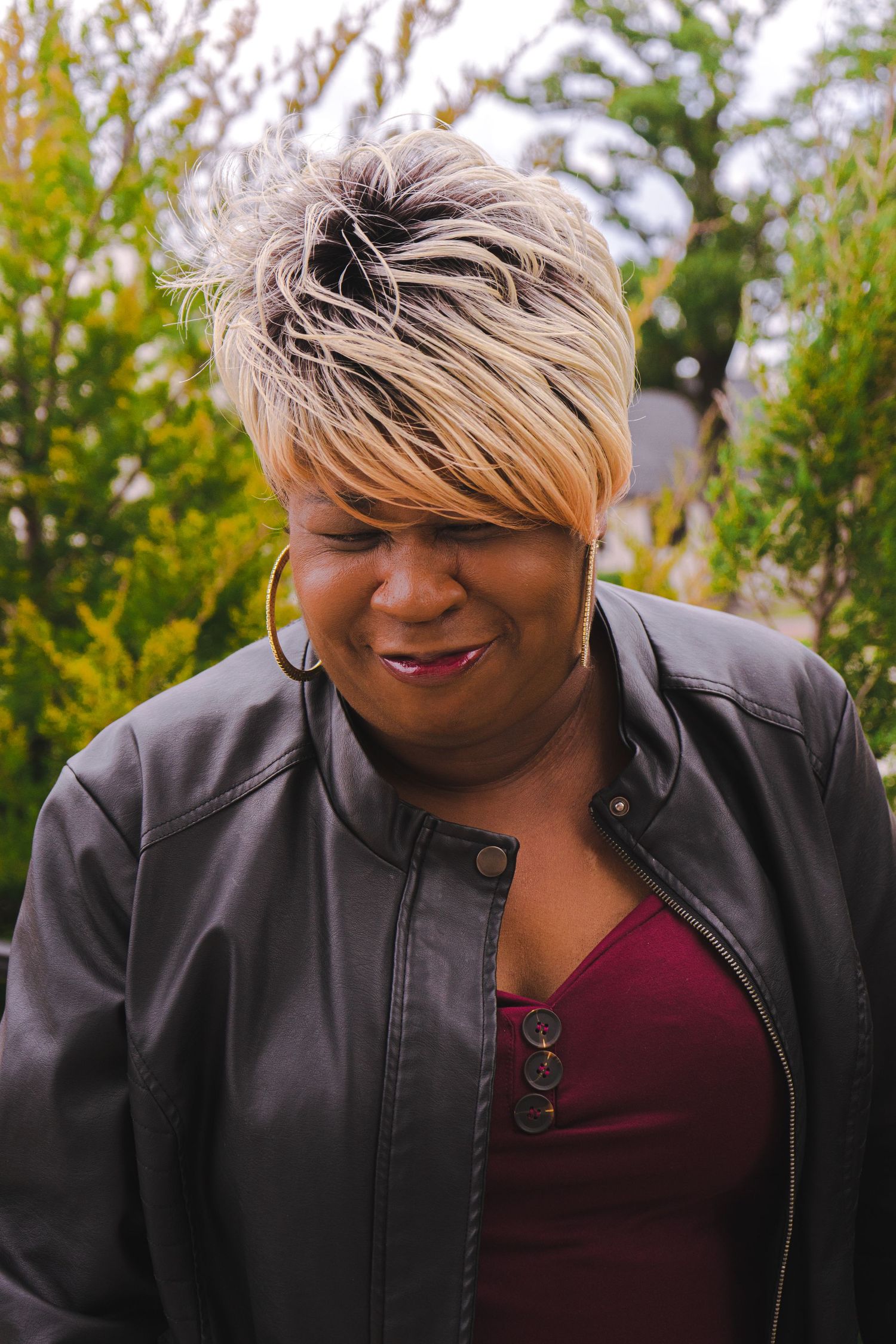 Professional portrait of person in black leather jacket and burgundy top smiling outdoors with flowering plants in background.