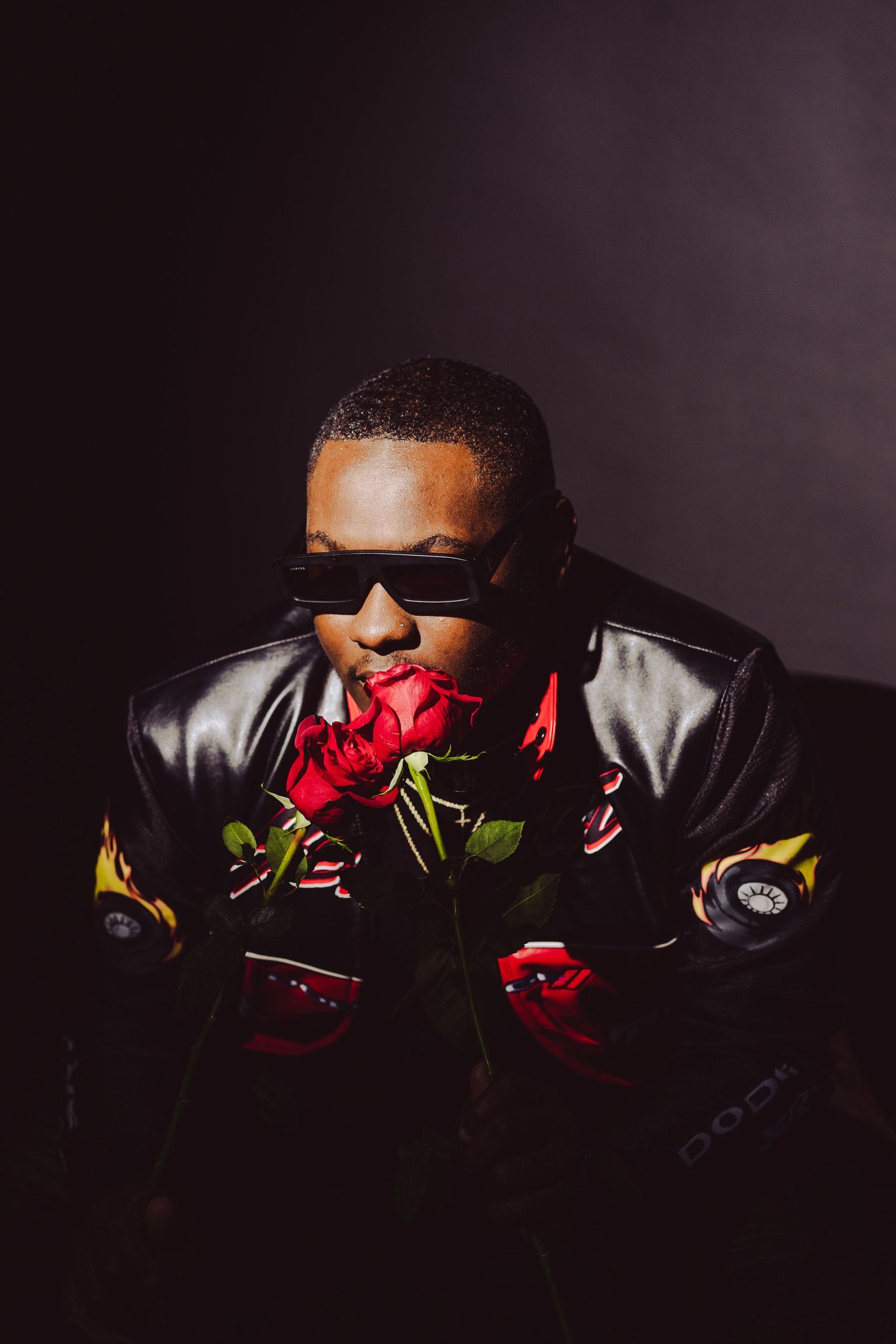 Dramatic portrait of person in sunglasses and leather jacket holding red roses in dark setting.