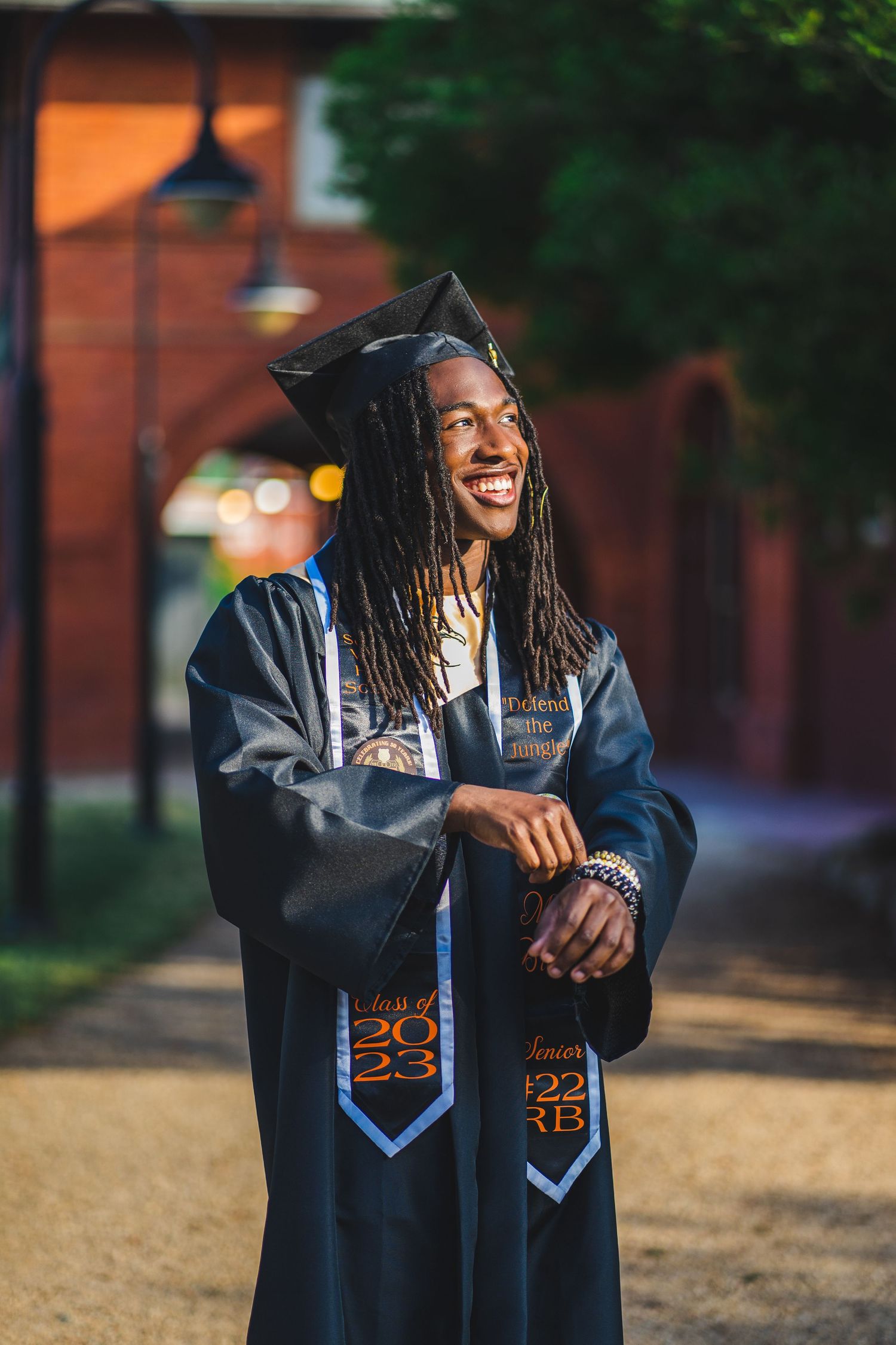 Graduate wearing a black academic gown with decorative stoles smiling joyfully on campus.