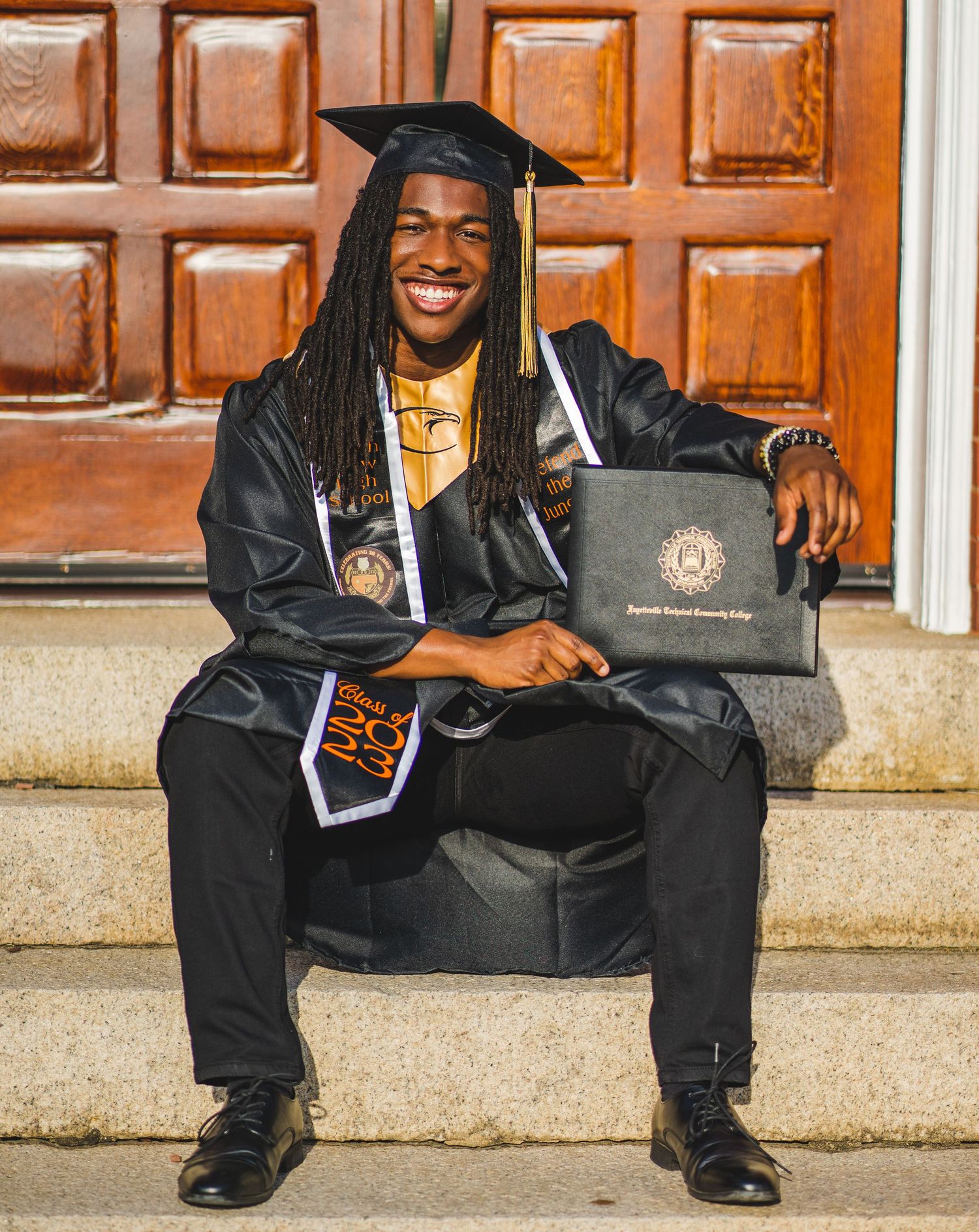 Graduate in cap and gown sits on stone steps with diploma and decorative stoles.