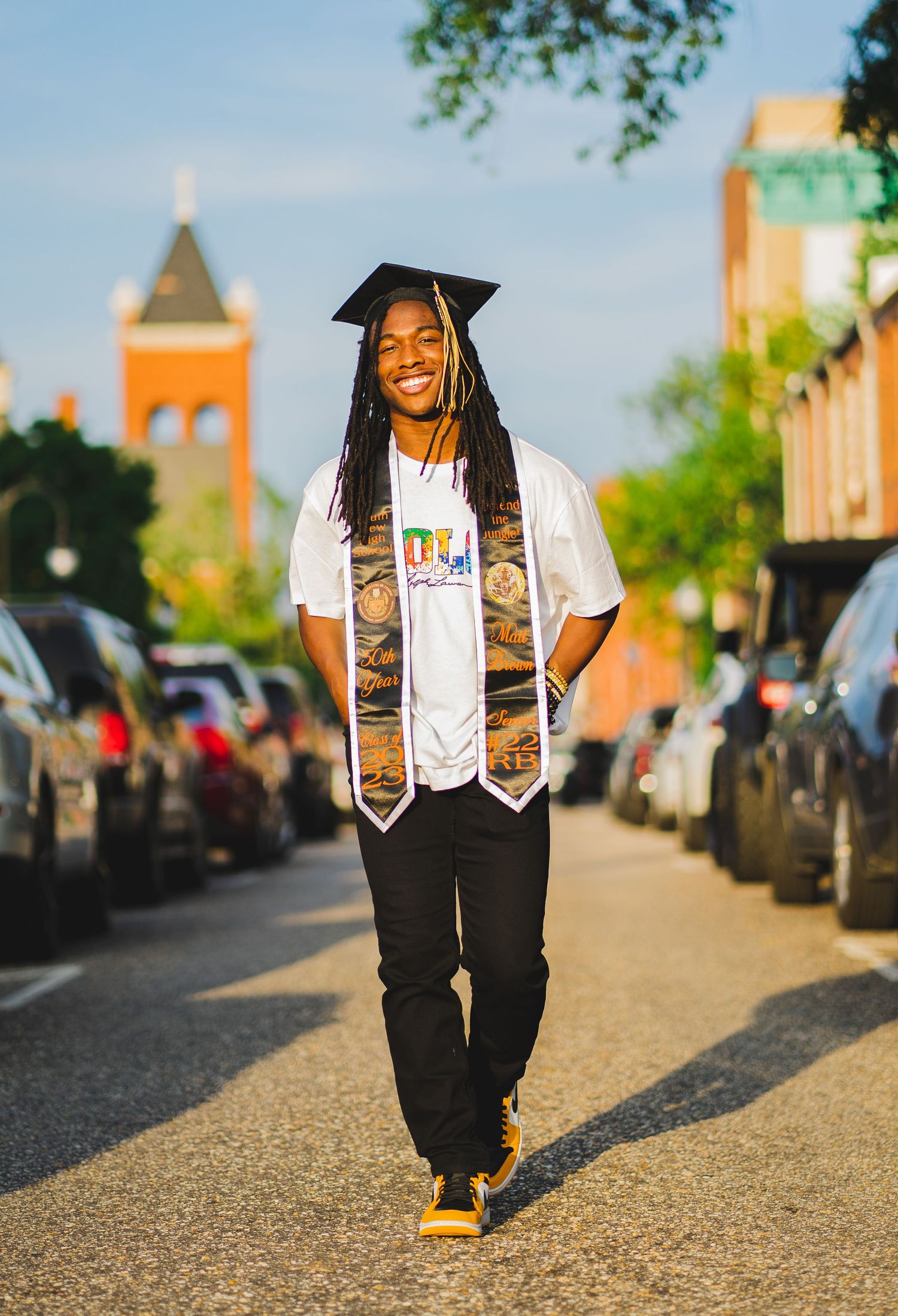 Graduate in cap and gown with stoles poses on a street with church tower in background.
