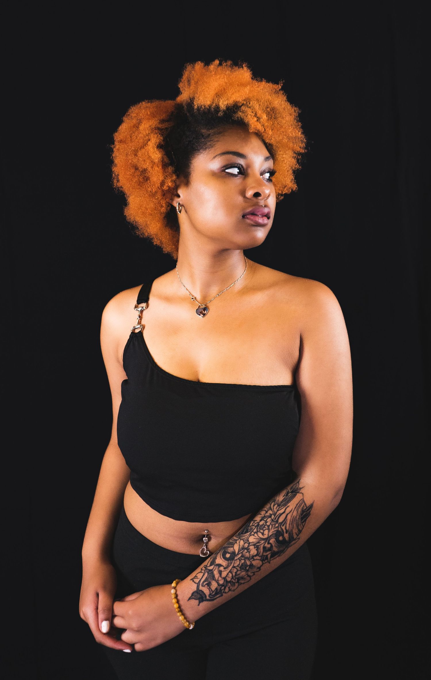Studio portrait with black background featuring person with orange natural hair wearing black crop top.