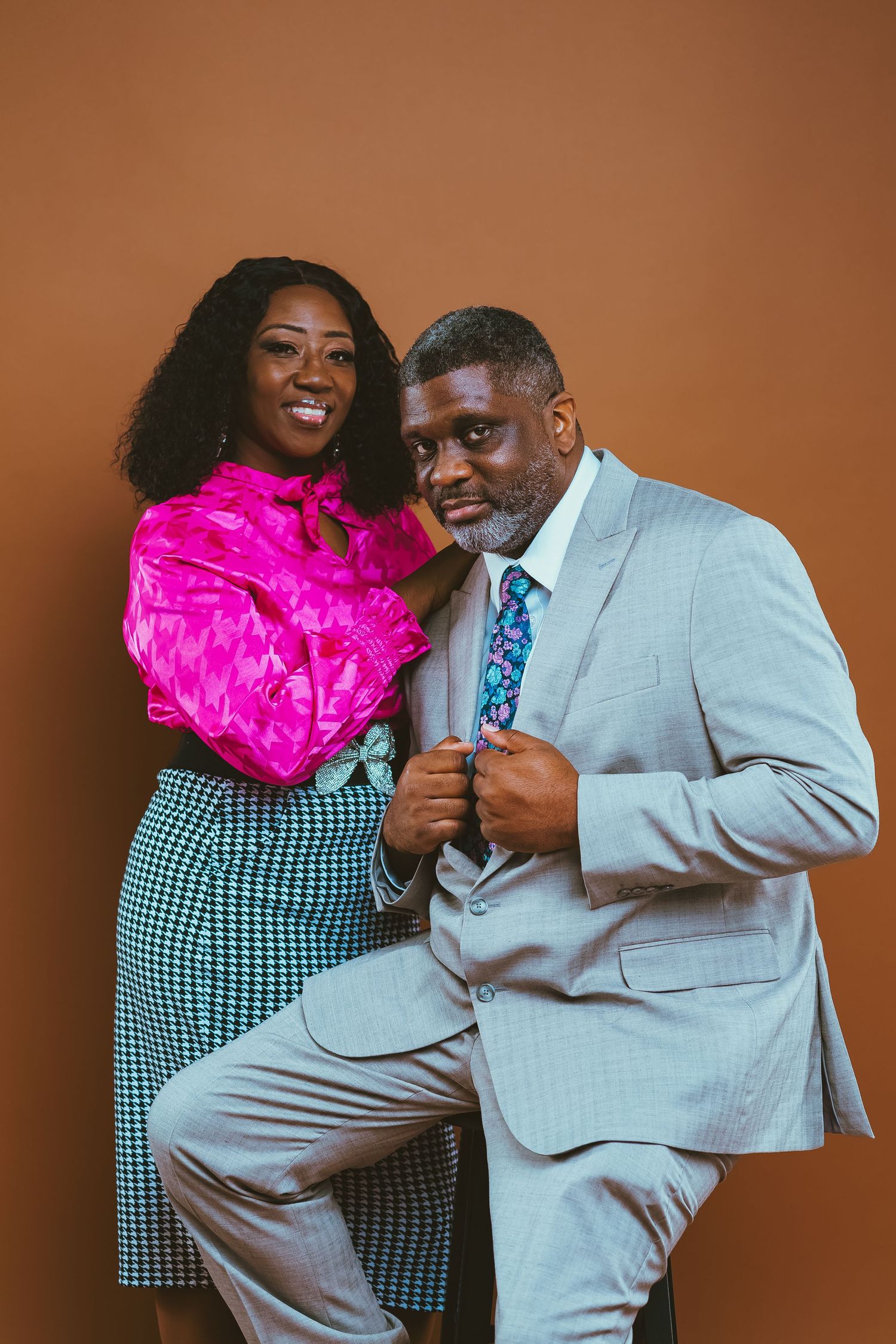 A stylish couple in pink and grey formal wear posing against a brown backdrop.