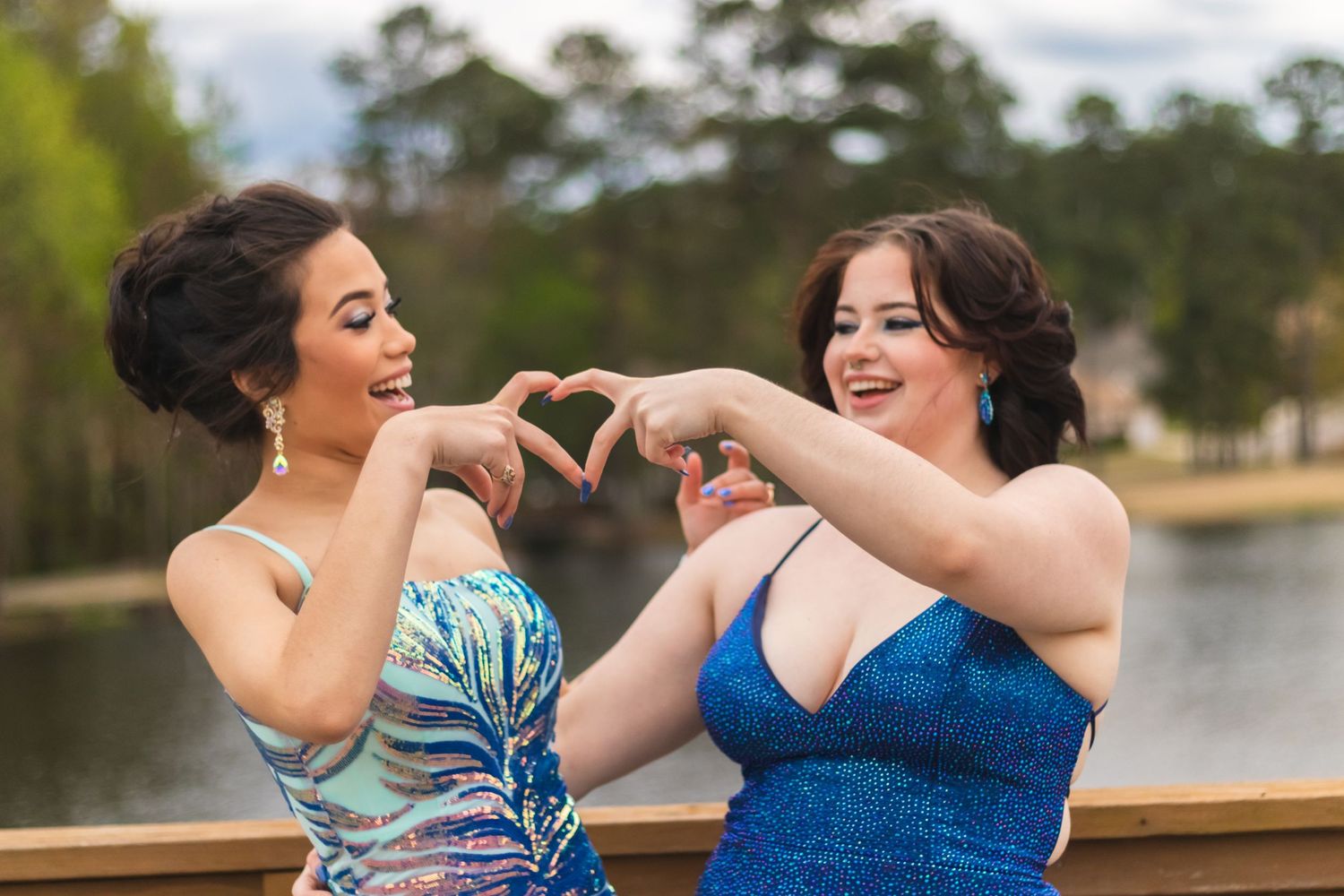 Two women in formal dresses making heart shape with hands on wooden deck.