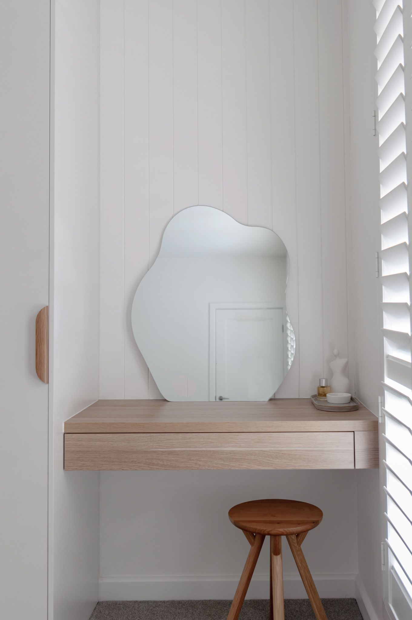 Minimalist vanity nook with organic-shaped mirror and wooden stool.