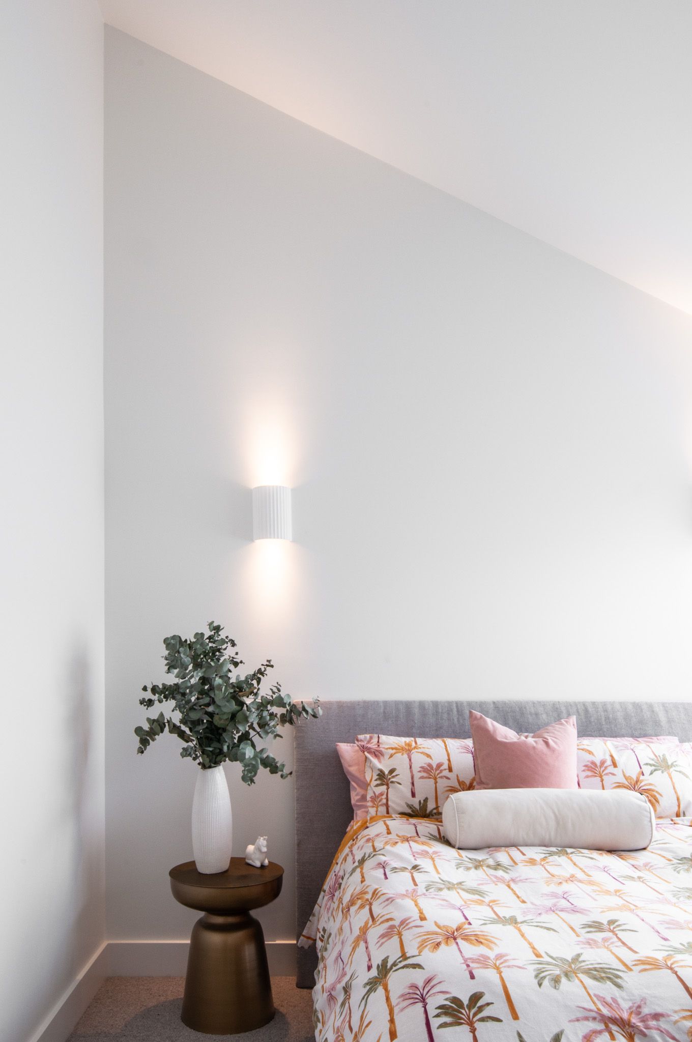 Bedroom with palm print bedding, pink accents, and modern wall sconce against white walls.