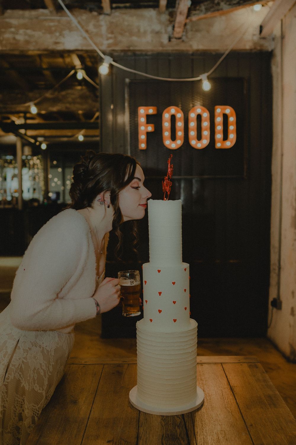 Bride blowing out candle on white wedding cake with heart details, holding beer, under a lit FOOD sign.