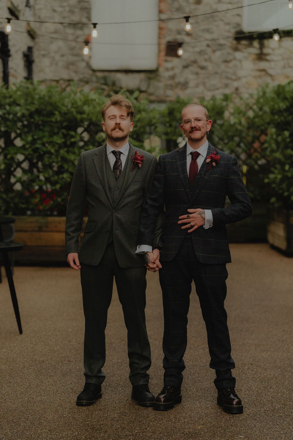 Two grooms in dark suits with red boutonnieres hold hands in a courtyard on their wedding day.