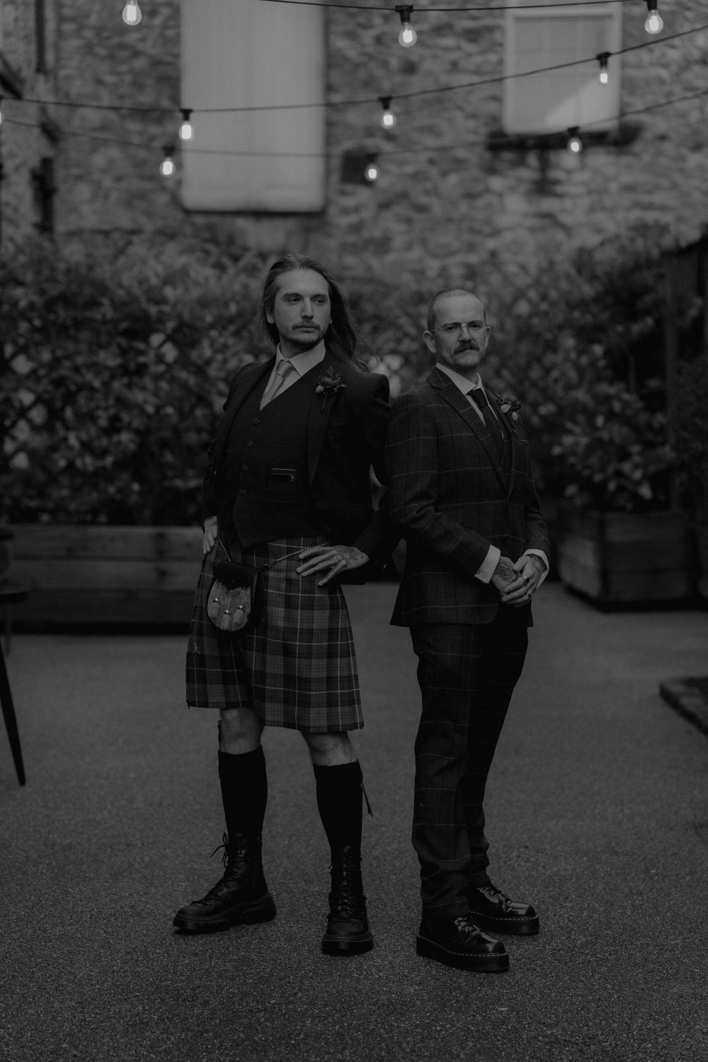 Two men pose in a courtyard, one wearing a tartan kilt and boots, the other in a suit, with string lights above.