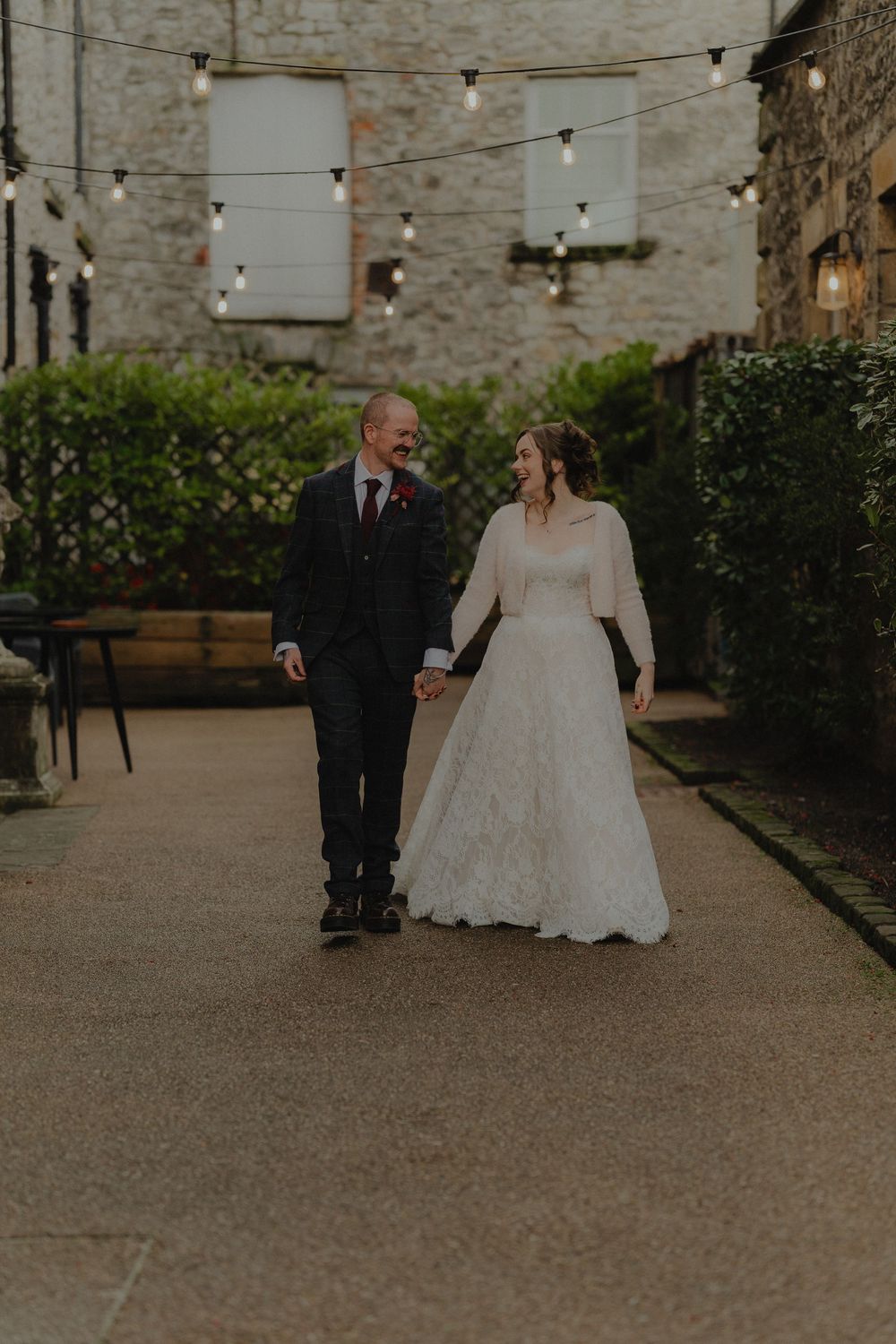 Bride and groom walk hand-in-hand through a stone courtyard adorned with string lights at their wedding.