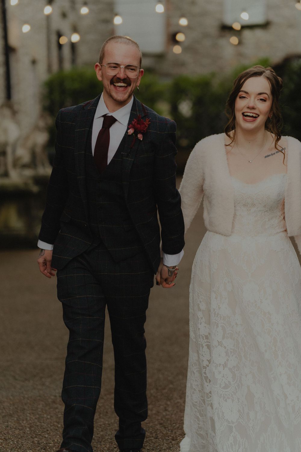 Laughing bride and groom in plaid suit walk hand in hand at outdoor wedding venue with string lights.