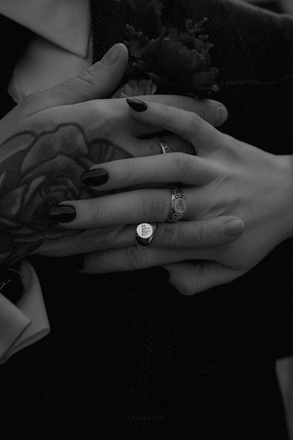 Black and white photo of two hands with tattoos, dark nails, and silver rings resting together.