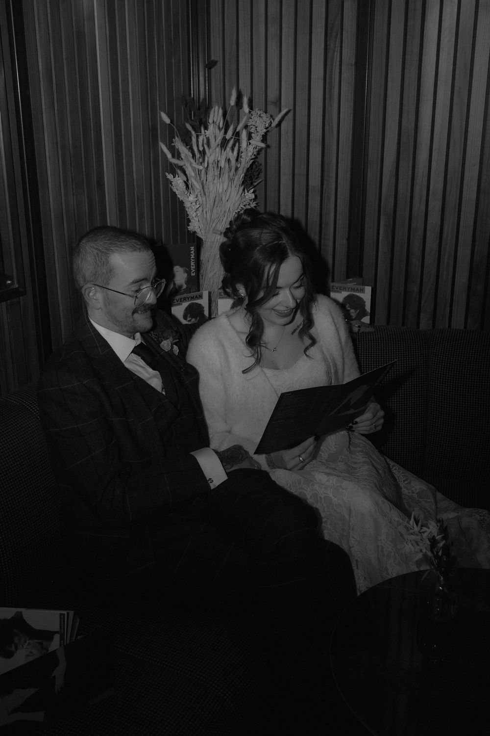 Black and white photo of a couple in wedding attire sitting on a sofa, smiling while reading a document together.
