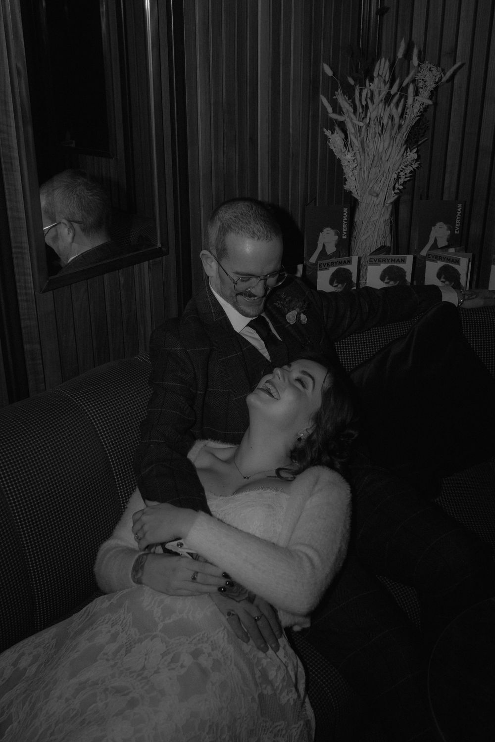 Black and white wedding photo of a groom embracing his laughing bride on a sofa, with decor visible behind them.