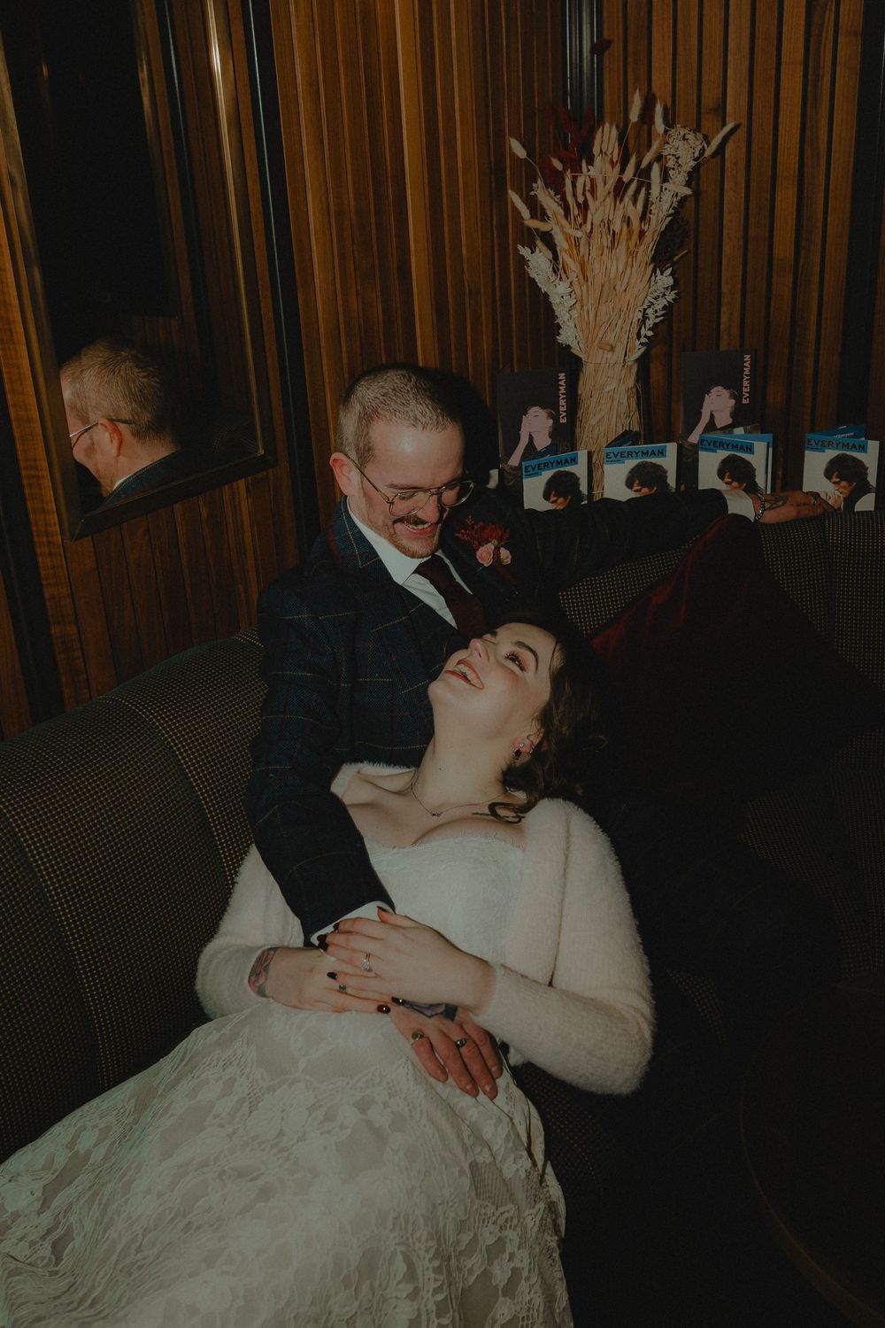 Bride in white lace dress laughs joyfully while groom holds her on a velvet sofa at wedding reception.