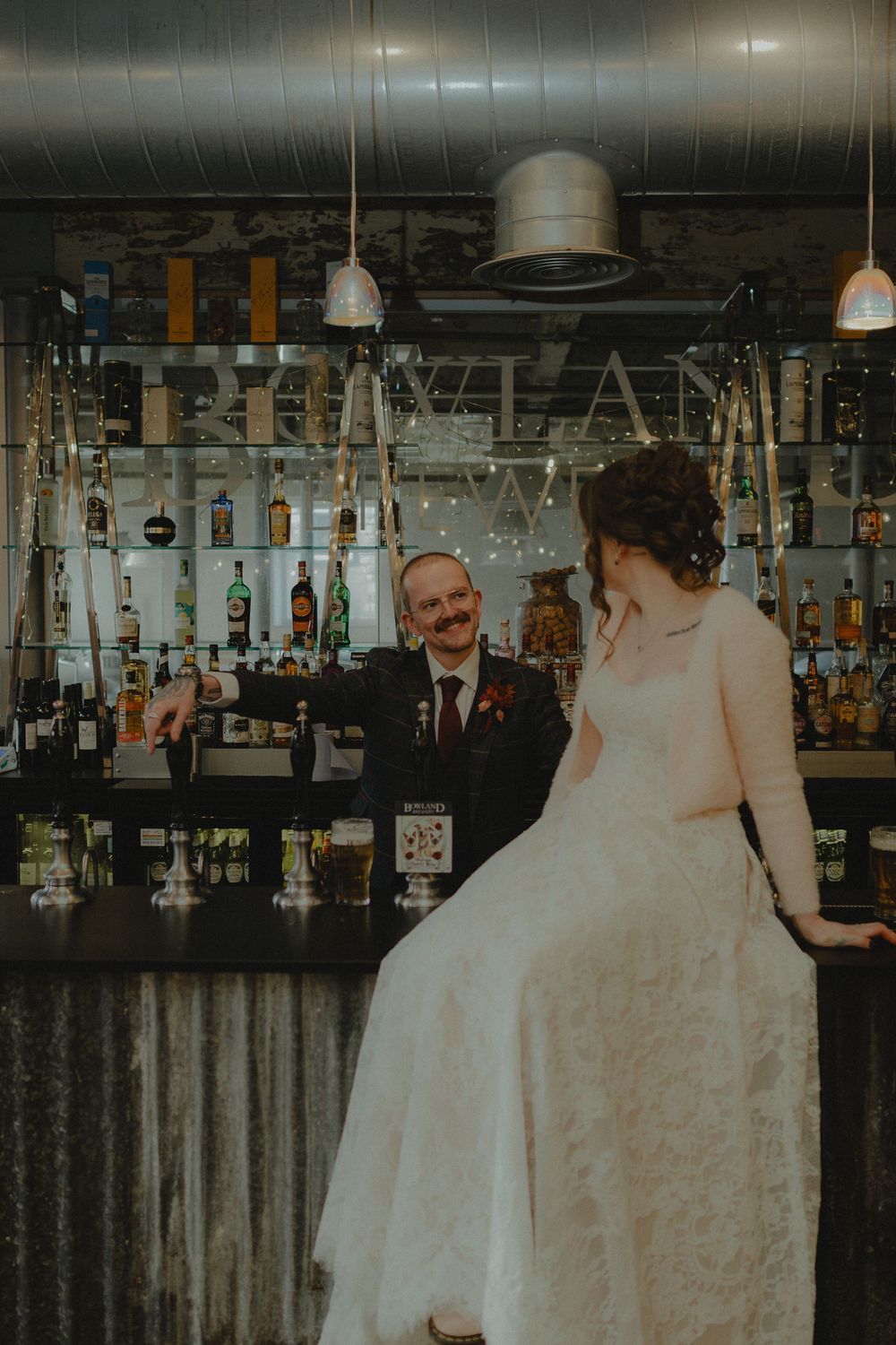 Bride in lace gown sits on bar counter as groom in suit pours a drink at a decorated wedding venue bar.