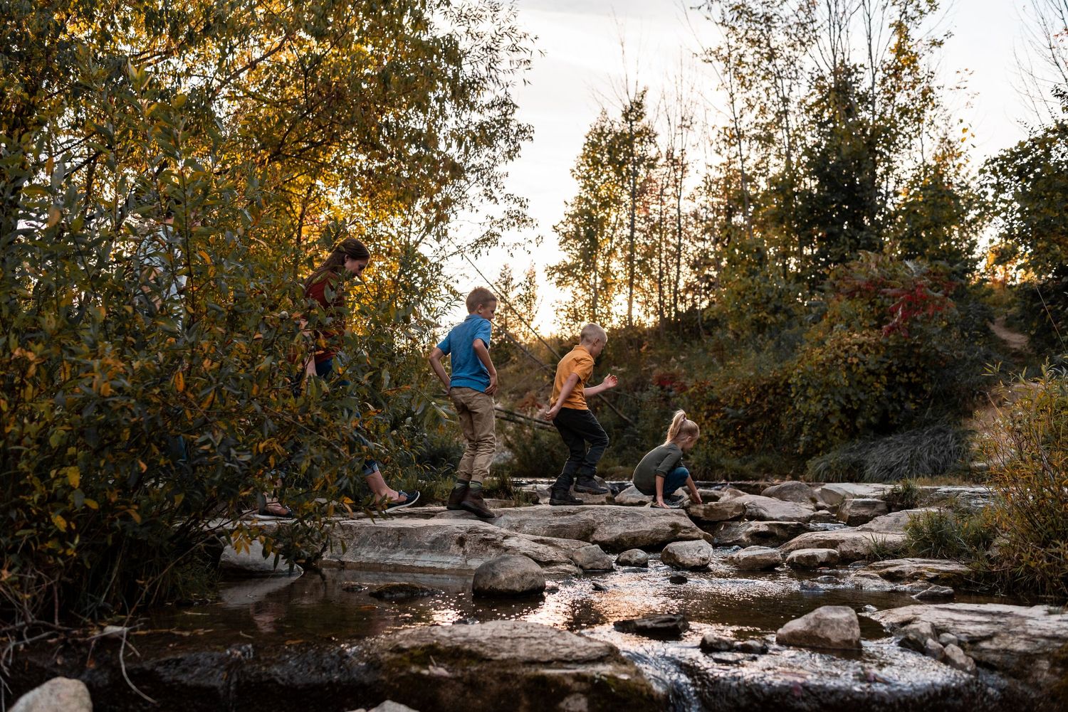 Children jumping across rocks in a stream at sunset with trees in the background.