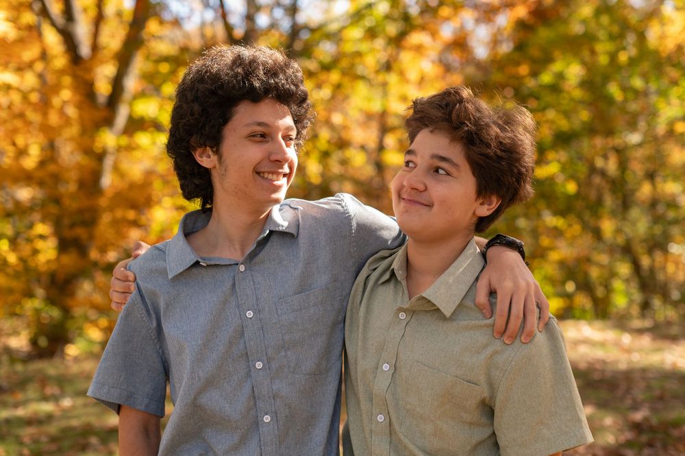 Two young people embrace while smiling together outdoors with vibrant autumn foliage in the background.