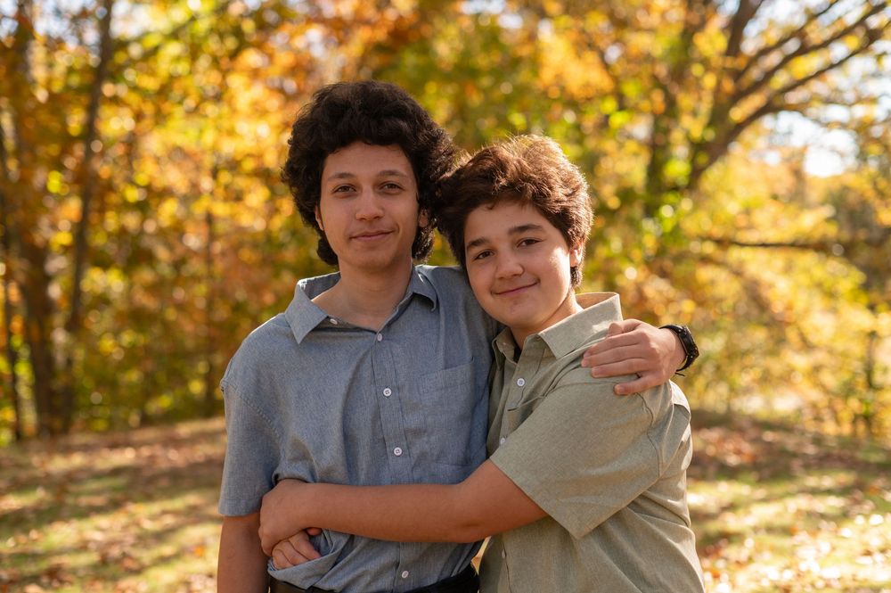 Two young people embrace while smiling outdoors on an autumn day with golden foliage in the background.