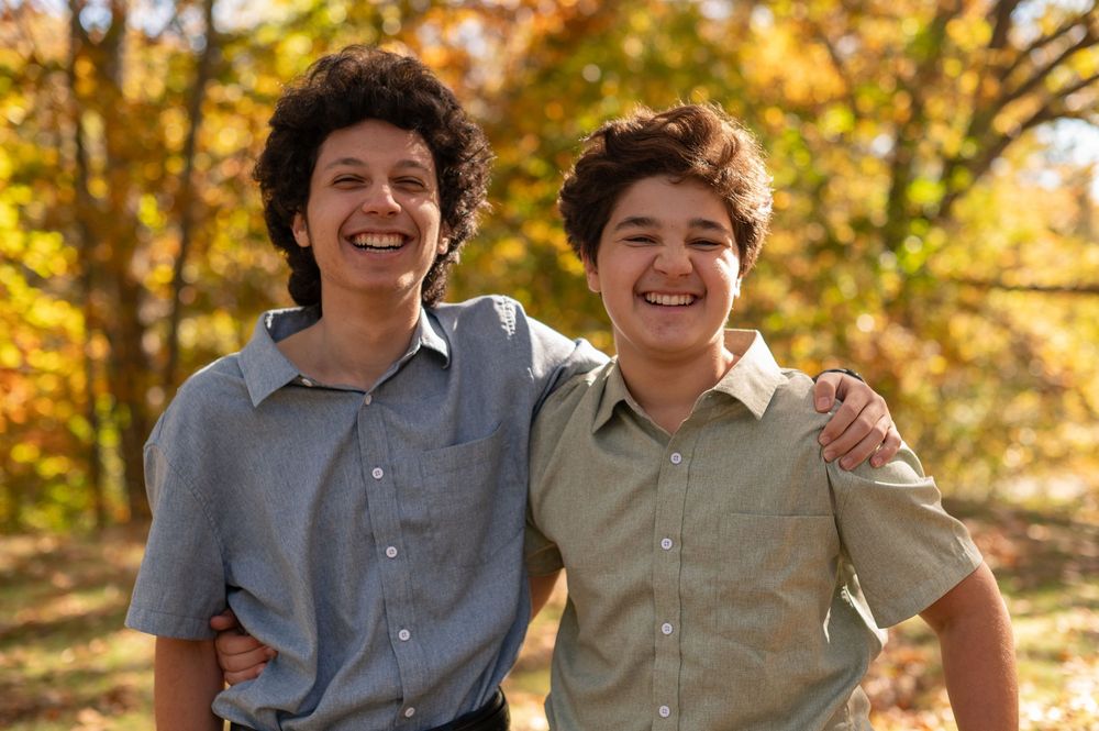 Two smiling siblings wearing button-down shirts embrace outdoors with autumn foliage in the background.