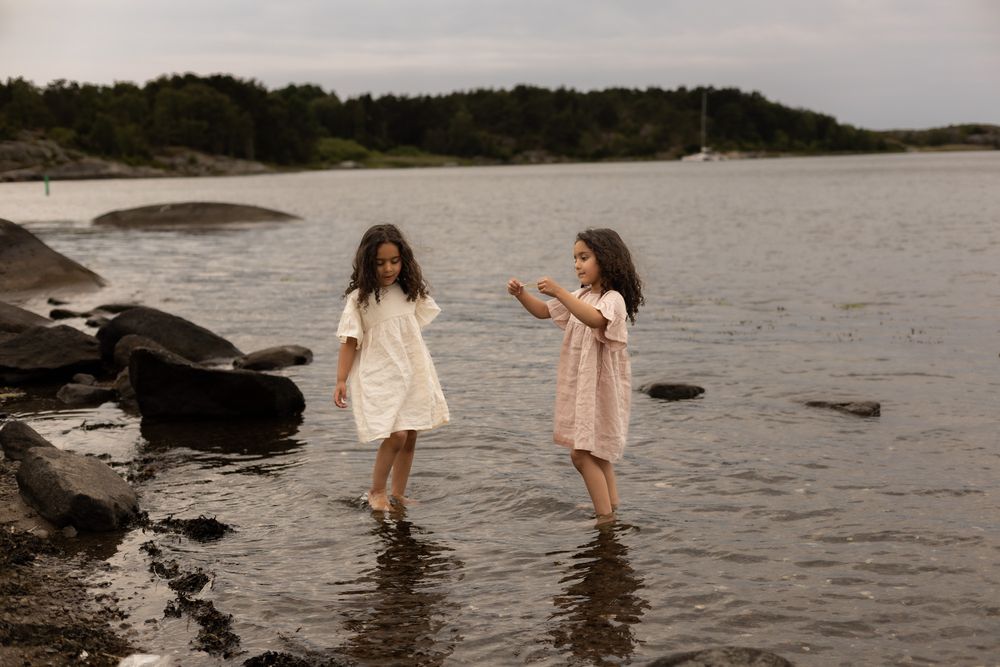 två barn som står i vattnet vid strandkanten. de har klänning på sig där den ena har en vit och den andra har en rosa