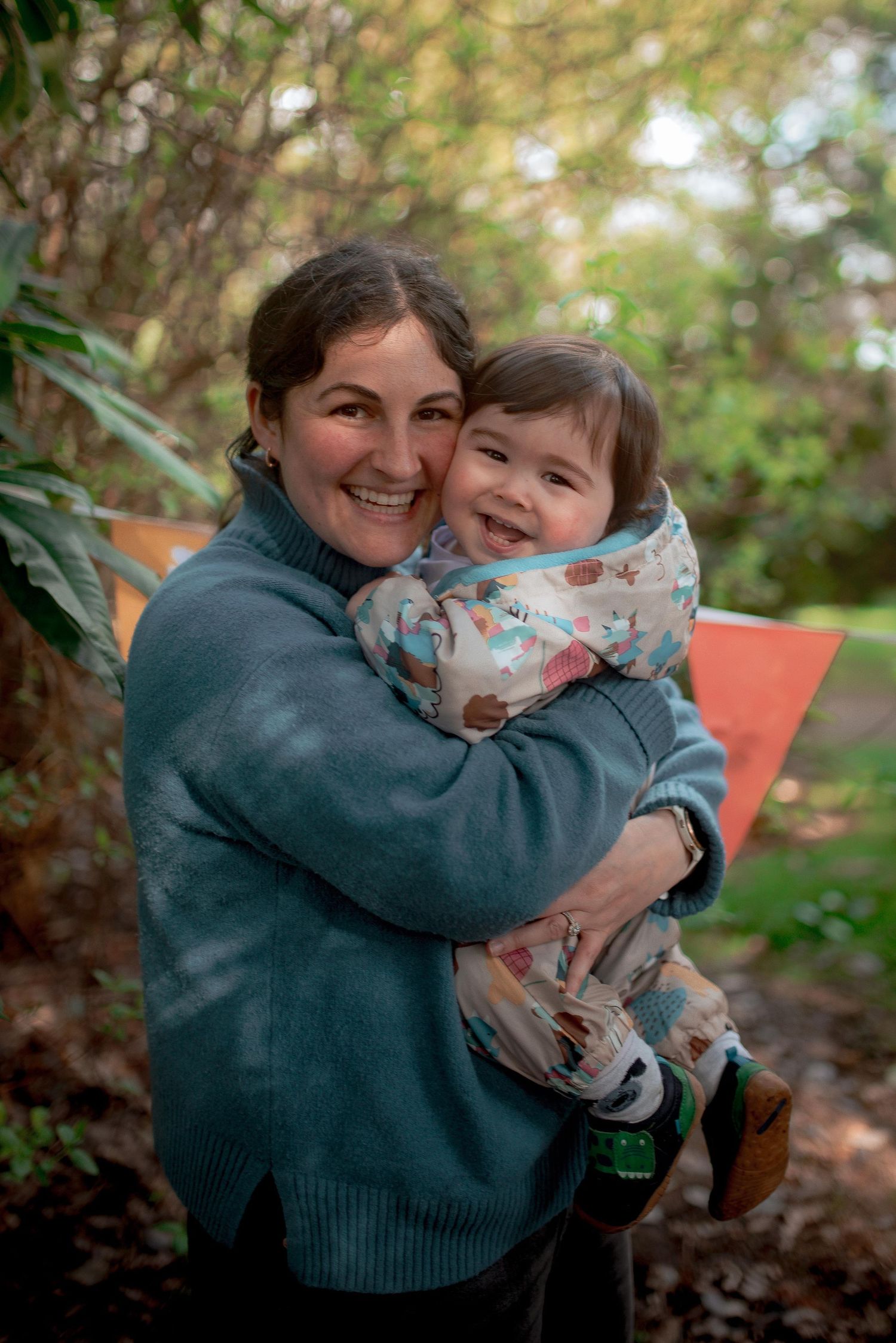 mum in blue and baby in a puddle suit hug and smile at the camera in dappled light