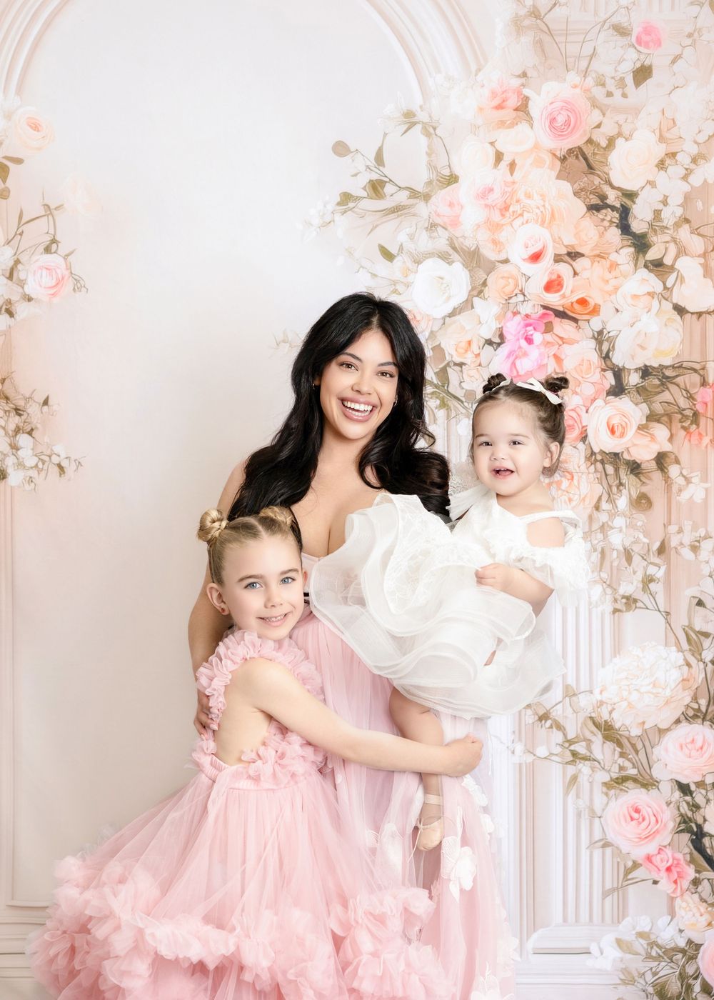 A smiling woman in white holds two young girls in pink and white dresses amid floral decor.