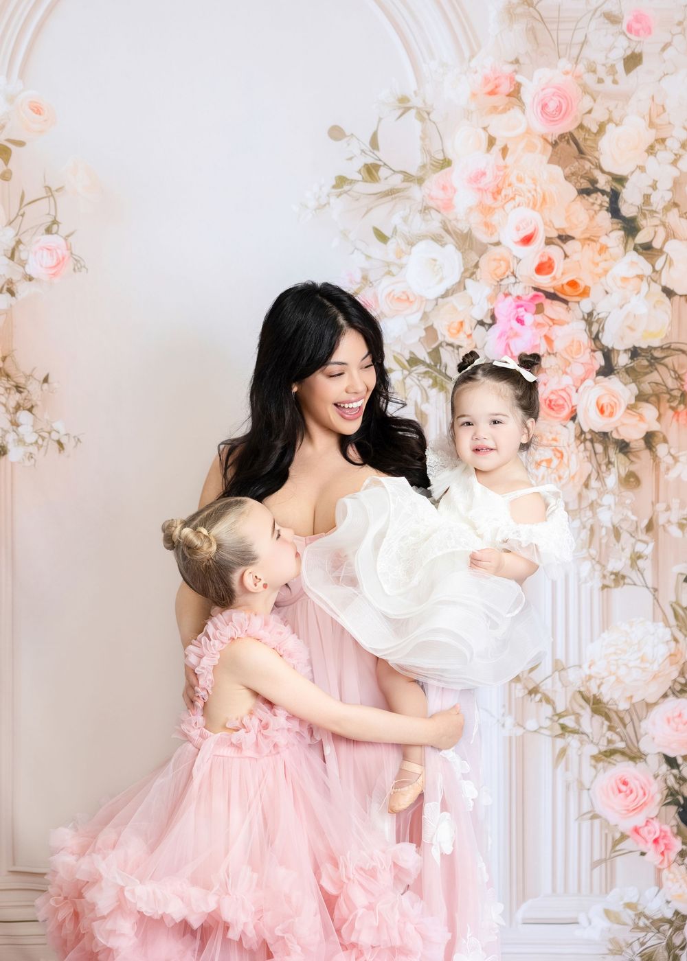 A woman in pink tulle gown holds two little girls in matching pink dresses, posing before a floral arch backdrop.