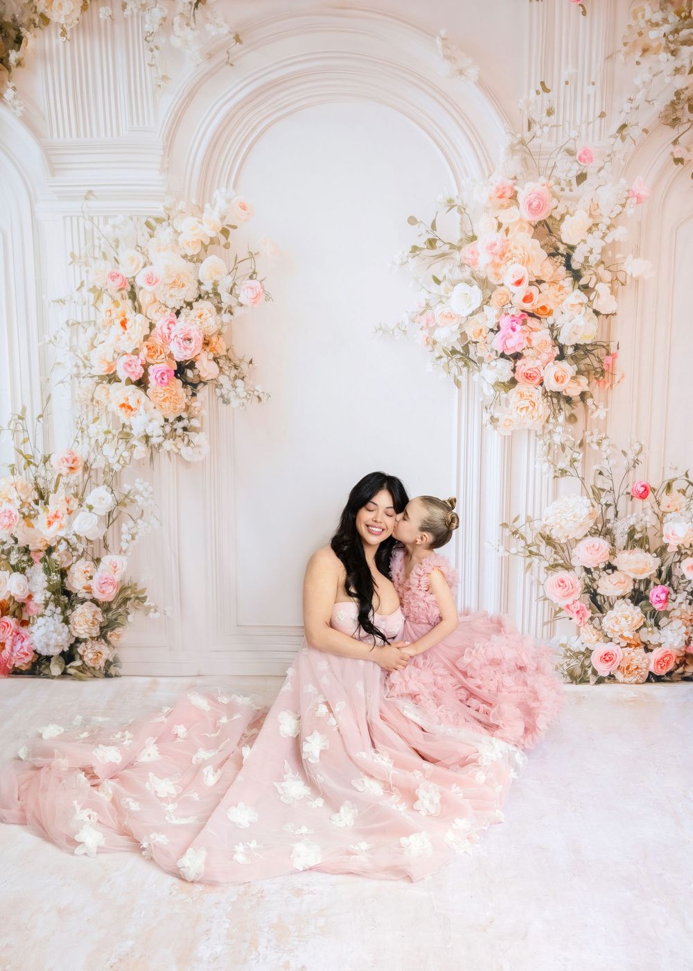 A mother and daughter in matching pink floral gowns sit together before an elegant floral arch backdrop.