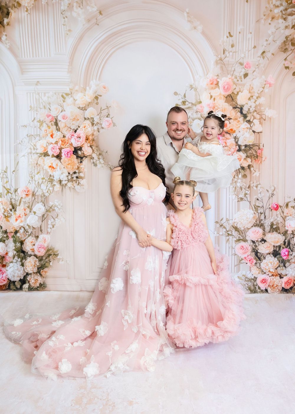 Family maternity photo with mom in pink floral gown, dad holding toddler, and daughter in pink dress, surrounded by flowers.