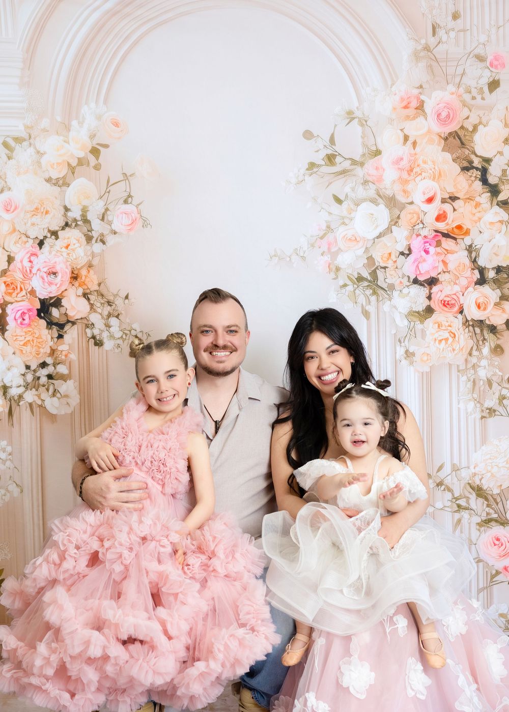 Family of four posing in front of floral arch, two girls in pink and white dresses, parents smiling in elegant studio setting.