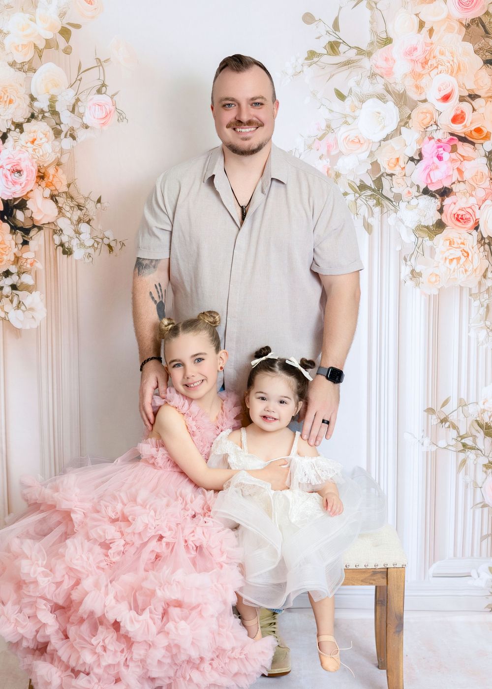 A man poses with two young girls in pink and white dresses in a floral studio setting.