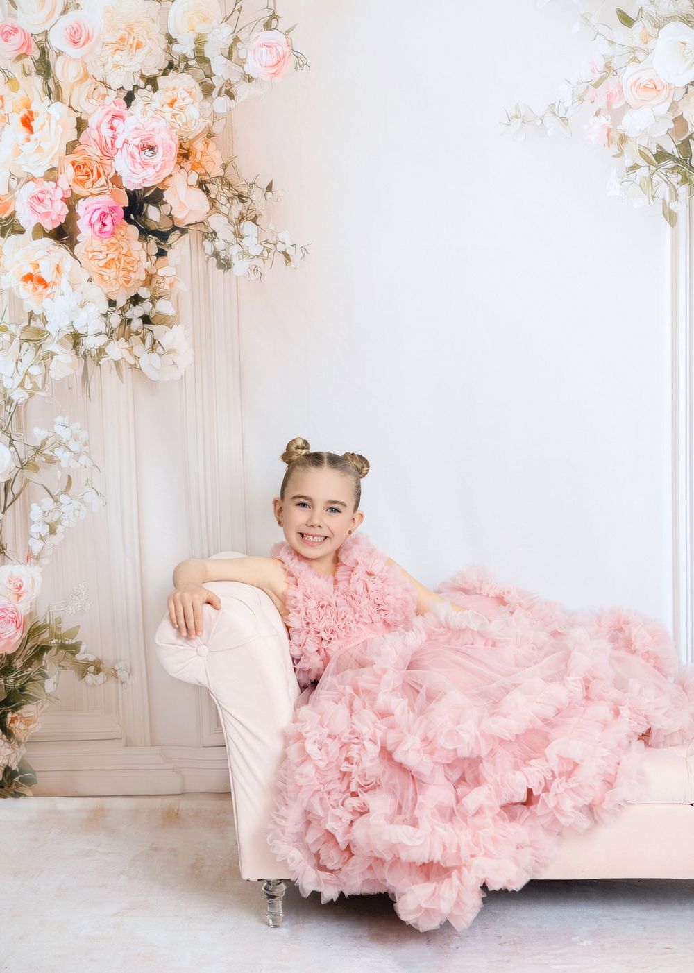 Young girl in pink ruffle gown poses on cream chaise lounge surrounded by pastel floral arrangements.
