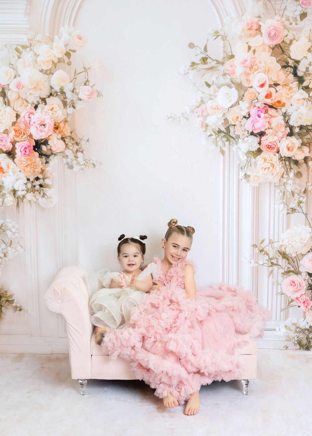 Two young girls in pink dresses pose on a small sofa surrounded by elegant floral arrangements in a bright studio.