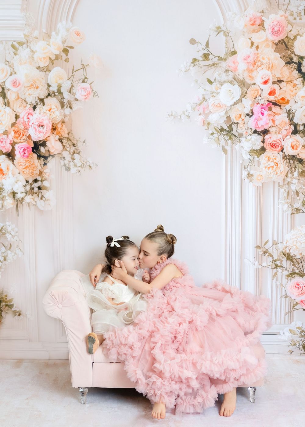Two young girls in pink and white dresses share a sweet kiss on a small sofa with floral decor.