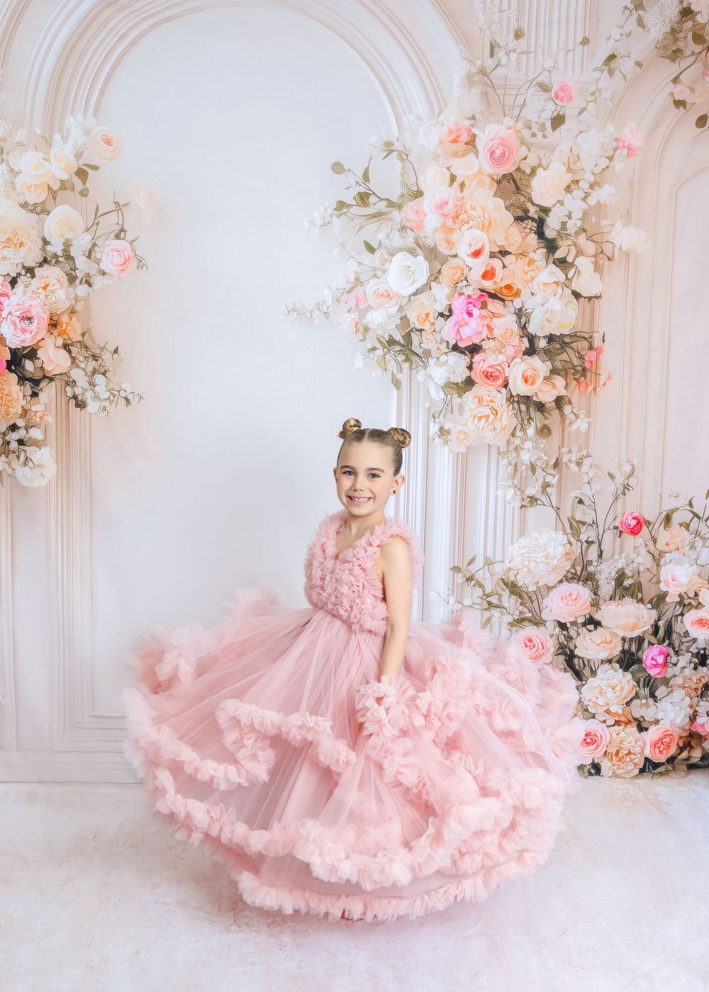 Young girl twirling in a pink ruffle tulle gown amid elegant floral arch decor with roses.