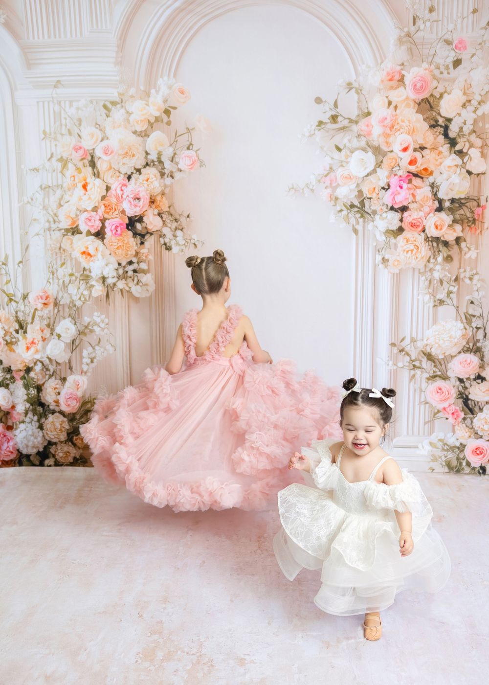Two young girls in pink and white ruffled dresses twirl and play among elegant floral arrangements.