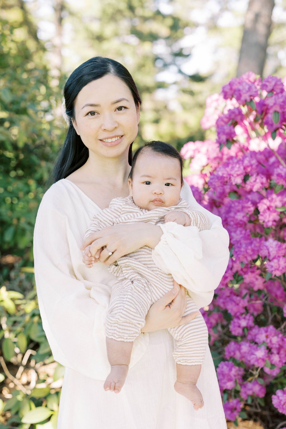 Smiling Asian mother in white dress holds baby in striped onesie near vibrant pink rhododendrons.