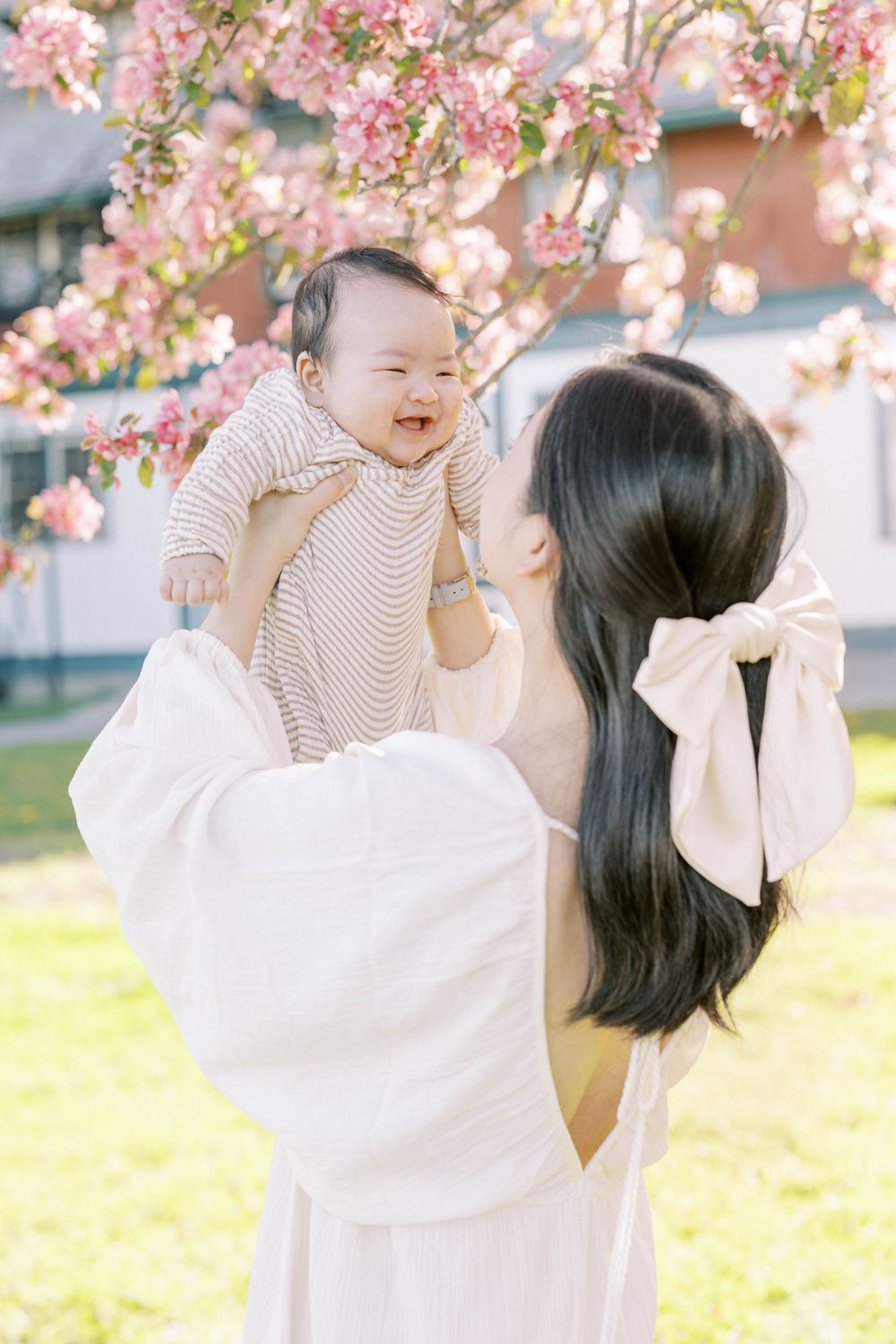 Mother lifting smiling baby girl under pink cherry blossom tree outdoors.