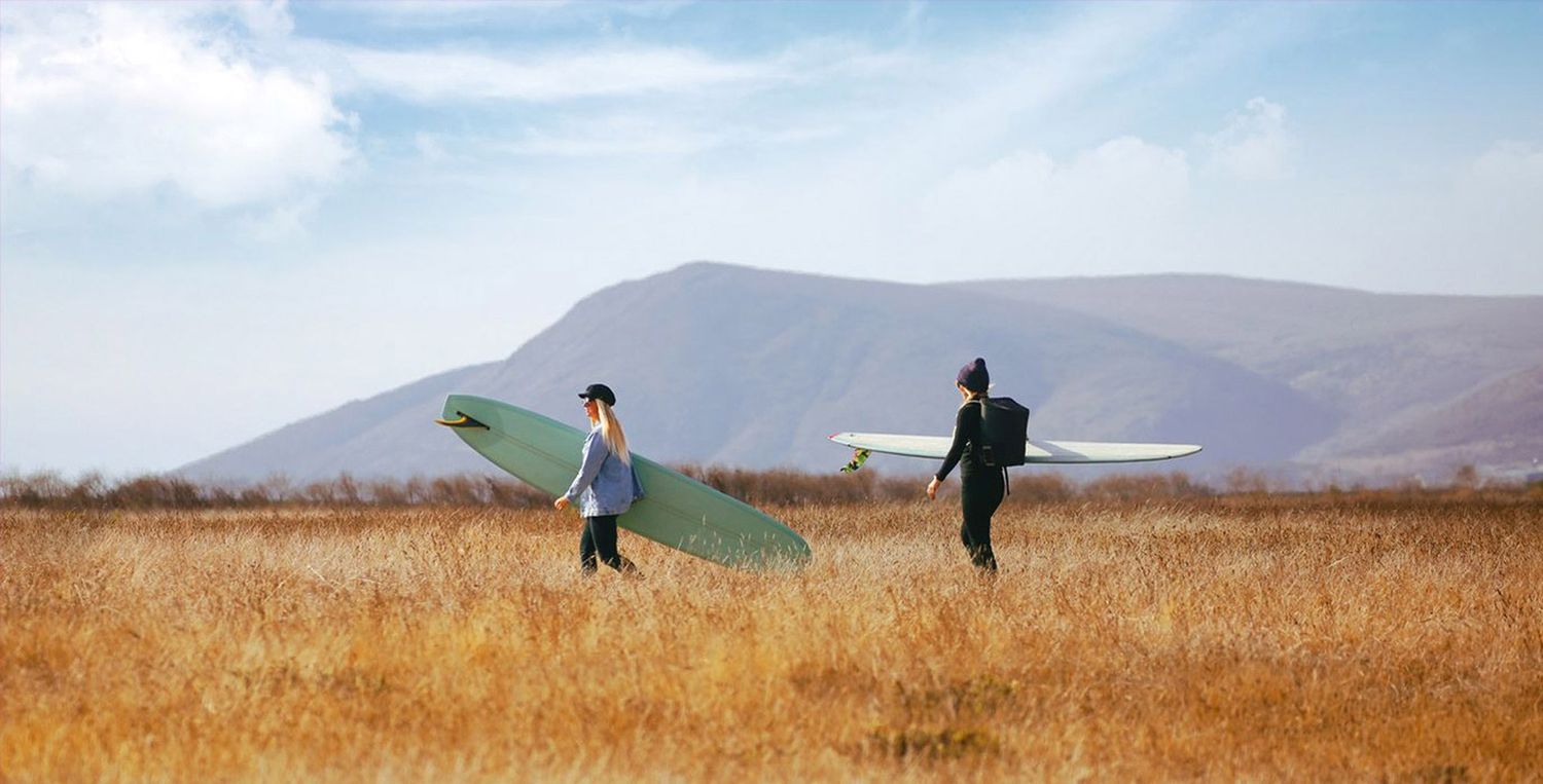 Two people walking through golden field carrying surfboards with mountains in background.