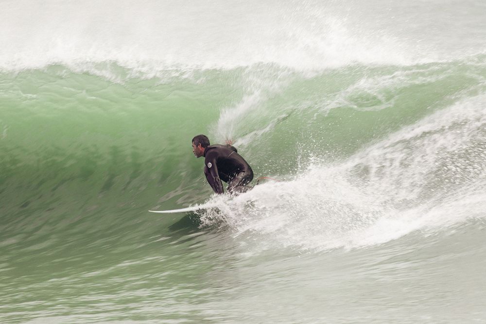 A surfer rides a large green wave in a wetsuit while creating a spray of white water behind their board.