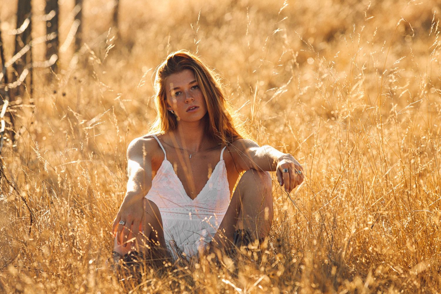 Woman in white dress sits thoughtfully in golden wheat field at sunset, backlit by warm sunlight.
