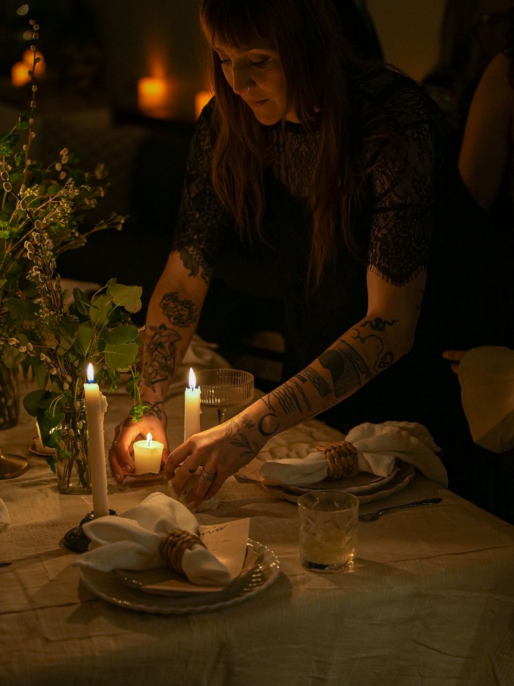 A candlelit table setting with someone in black lace arranging table decor among glowing candles and green foliage.