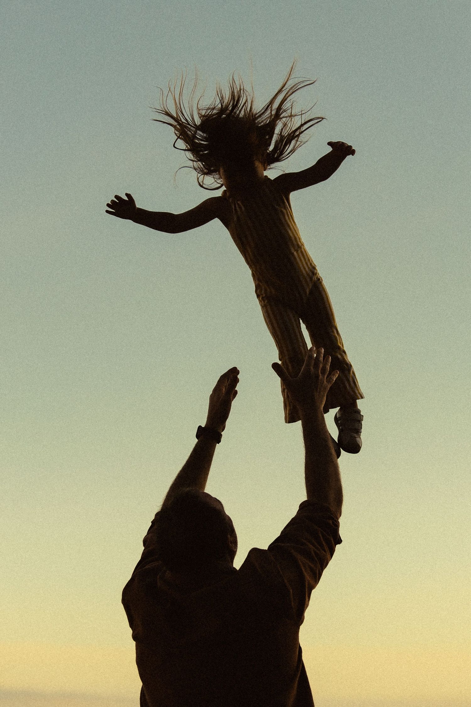 Silhouette of two figures against sunset sky with one person tossing another person joyfully into the air.